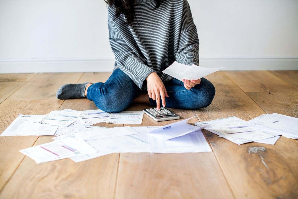 A woman sitting on the floor surrounded by sheets of paper using a calculator.