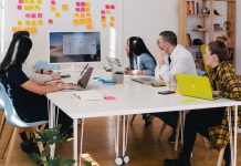 The Biggest Pros and Cons of a Four-Day Work Week Photo of a group of people having a meeting in an office and looking at a screen in front of a white table with post its on it as a featured image for a post about the pros and cons of a four-day work week