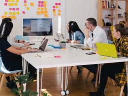 The Biggest Pros and Cons of a Four-Day Work Week Photo of a group of people having a meeting in an office and looking at a screen in front of a white table with post its on it as a featured image for a post about the pros and cons of a four-day work week