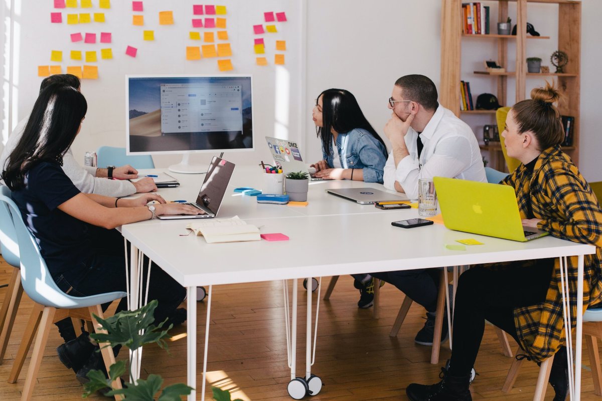 Photo of a group of people having a meeting in an office and looking at a screen in front of a white table with post its on it as a featured image for a post about the pros and cons of a four-day work week